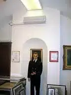 Mannequin in formal suit displayed in historic mansion gallery room with wall-mounted HVAC unit and framed artwork