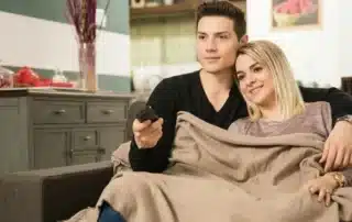 Man and woman sharing blanket on couch watching TV together in comfortably heated home