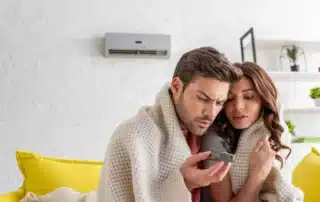 Couple wrapped together in knit blanket looking at thermostat remote control feeling cold indoors with mini-split unit visible on white brick wall background