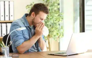 Man in denim shirt sneezing into tissue at office desk with laptop suffering from indoor allergies and poor air quality