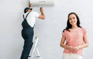 HVAC technician in overalls on ladder installing wall-mounted mini-split unit while smiling female homeowner in striped shirt stands nearby on white brick wall background