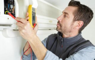 Professional HVAC technician in vest using yellow multimeter with red probes to diagnose and repair wall-mounted HVAC unit electrical components