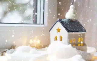 Miniature ceramic house wearing knit beanie hat surrounded by snow and warm glowing lights on windowsill during heavy snowstorm symbolizing home furnace preparedness