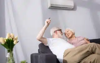 Happy senior couple relaxing on sofa using remote control to adjust wall-mounted mini-split air conditioner in comfortable home