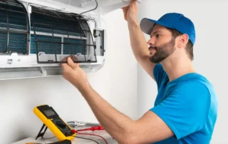 HVAC technician in blue shirt and cap using yellow multimeter to test and service wall-mounted mini-split air handler during winter maintenance inspection