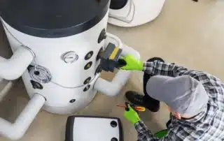 HVAC technician in plaid shirt and green gloves kneeling on floor servicing large water heater tank with tools during heating system repair in Connecticut