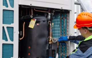 HVAC technician wearing orange hard hat and blue gloves inspecting commercial refrigeration unit with copper pipes and coils in Connecticut